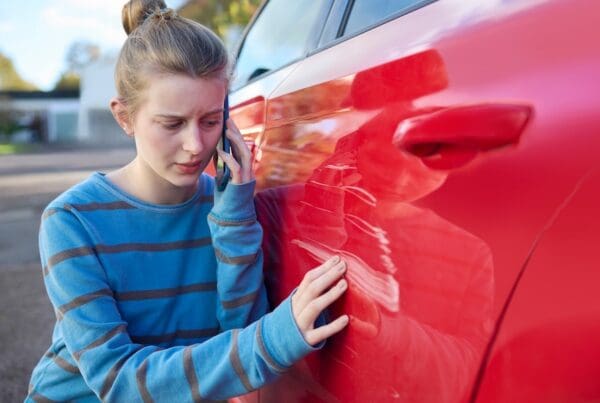 Young Worried Teenage Female Driver With Damaged Car After Accident Calling Insurance Company On Mobile Phone